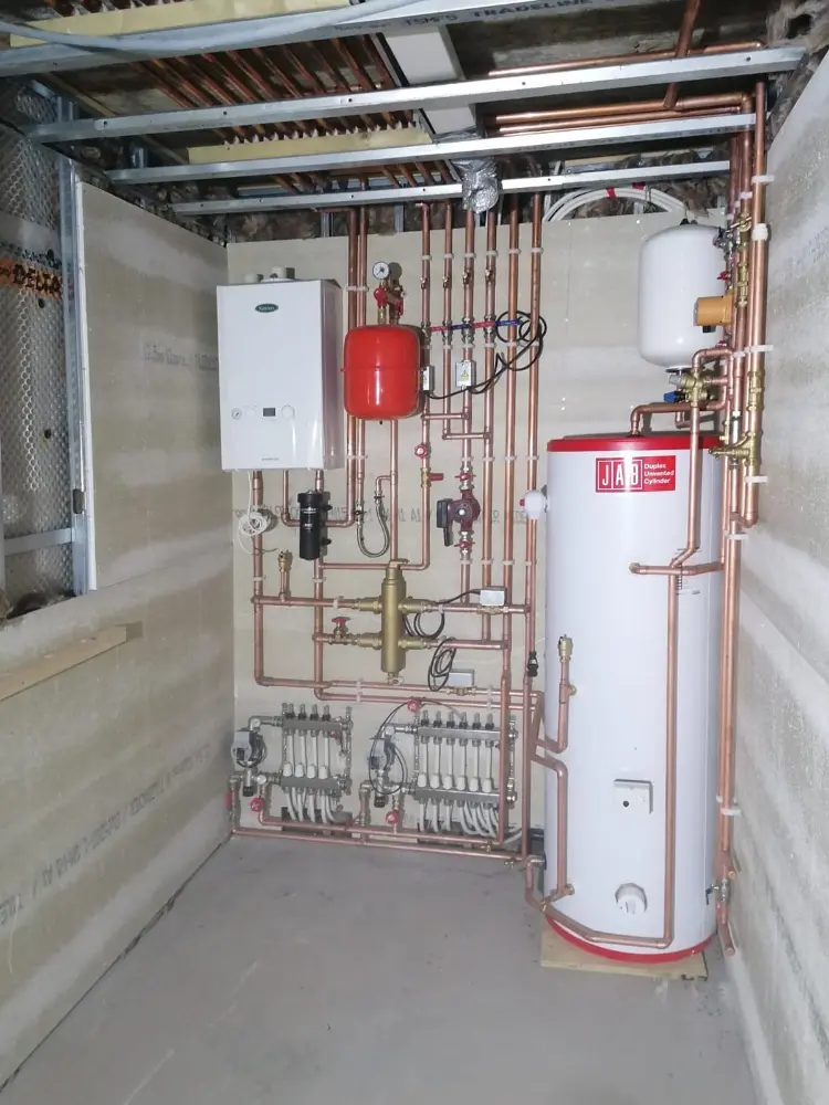 Kotłownia z podgrzewaczem wody, różnymi rurami, czerwonym zbiornikiem wyrównawczym i zaworami regulacyjnymi zamontowanymi na ścianie.