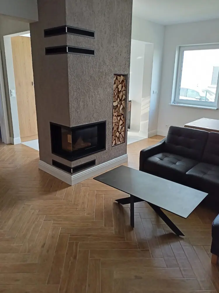 Nowoczesny salon z kominkiem, schowkiem na drewno, czarną kanapą i minimalistycznym stolikiem kawowym na parkiecie.