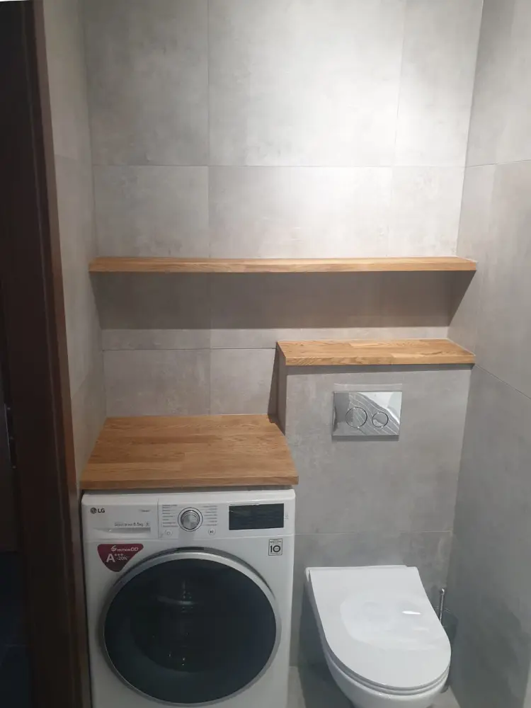 Kompaktowa łazienka z pralką, toaletą podwieszaną na ścianie i drewnianymi półkami nad i obok toalety.