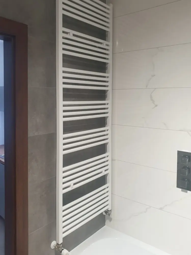 Wysoki, biały grzejnik na ręczniki zamontowany na kafelkowej ścianie łazienki.