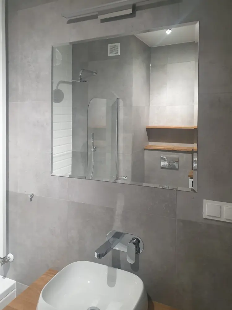 Nowoczesna łazienka z dużym lustrem, umywalką ścienną, baterią i widoczną kabiną prysznicową w odcieniach szarości.