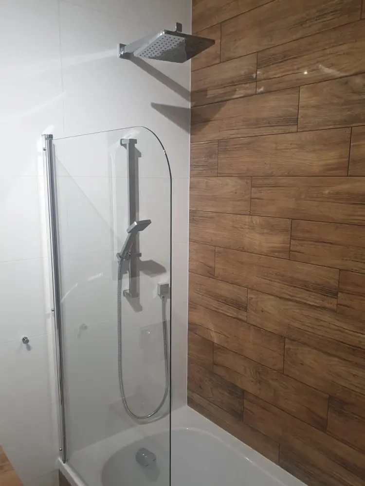 Nowoczesny prysznic ze szklaną ścianką, kwadratową słuchawką prysznicową i ścianą wyłożoną płytkami imitującymi drewno.